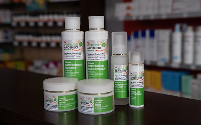 Sechs Flaschen und Tiegel mit Hautpflegeprodukten, die mit „Laurentius Apotheke Imst / Pitztal Apotheke“ beschriftet sind, stehen auf einer dunklen Theke, im Hintergrund sind verschwommene Regale zu sehen.
