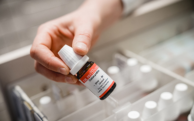 Eine Hand, die eine kleine braune Glasmedizinflasche mit der Aufschrift „Dr. Reckeweg“ aus der Laurentius Apotheke Imst / Pitztal Apotheke hält, über einer Schublade mit mehreren ähnlichen Flaschen.