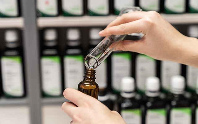 In der Laurentius Apotheke Imst / Pitztal Apotheke füllt eine Person Flüssigkeit aus einem Messzylinder in eine braune Glasflasche, im Hintergrund sind Regale mit beschrifteten Flaschen zu sehen.