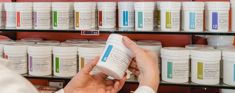 Eine Person hält in der Laurentius Apotheke Imst / Pitztal Apotheke ein weißes Plastikglas mit einem Aufkleber mit der Nummer 8 in der Hand, vor Regalen, die mit ähnlichen weißen Gläsern gefüllt sind, die jeweils verschiedenfarbige Nummernaufkleber tragen.