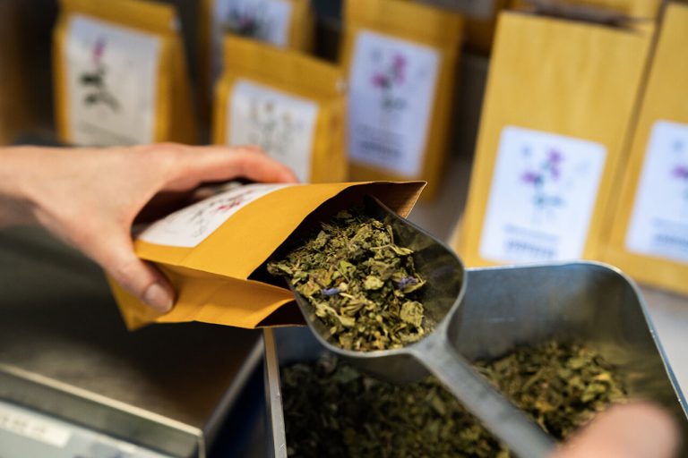 Eine Hand füllt mit einer Metallschaufel eine gelbe Papiertüte der „Laurentius Apotheke Imst / Pitztal Apotheke“ mit getrockneten Kräuterblättern; im Hintergrund sind ähnlich beschriftete Tüten zu sehen.