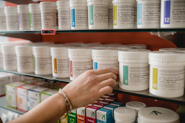 Eine Person greift nach einem weißen Plastikbehälter mit einem nummerierten Etikett in einem Regal der Laurentius Apotheke Imst / Pitztal Apotheke, in dem ähnliche Behälter und Medikamentenschachteln aufbewahrt werden.