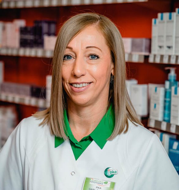 Eine Frau in Apothekenuniform steht lächelnd vor Regalen, die mit verschiedenen Arzneimitteln gefüllt sind, in der Laurentius Apotheke Imst / Pitztal Apotheke.