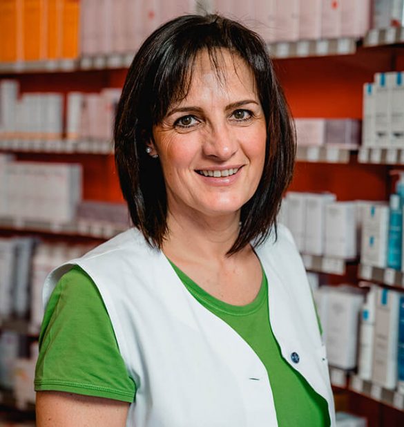 Eine Frau in einem grünen Hemd und einer weißen Weste steht vor Regalen voller verpackter Produkte in der Laurentius Apotheke Imst / Pitztal Apotheke und lächelt.