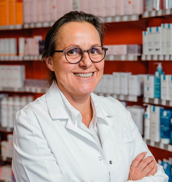 Ein Apotheker in einem weißen Laborkittel steht mit verschränkten Armen und einem Lächeln vor Regalen, die mit verschiedenen Arzneimitteln gefüllt sind, in der Laurentius Apotheke Imst / Pitztal Apotheke.