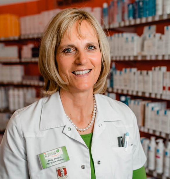 Eine Frau in einem weißen Apothekenkittel steht lächelnd vor den mit Produkten gefüllten Regalen der Laurentius Apotheke Imst / Pitztal Apotheke; sie trägt ein Namensschild und eine Perlenkette.