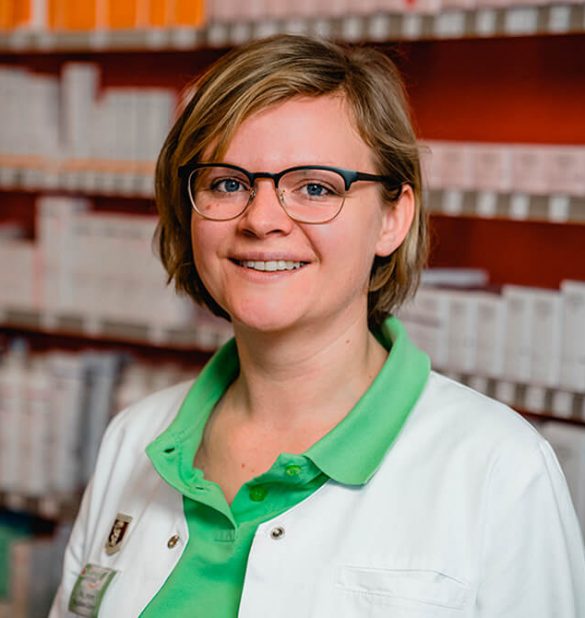 Eine Frau mit Brille und einem weißen Kittel über einem grünen Hemd steht vor Regalen, die mit Kartons gefüllt sind, in der Laurentius Apotheke Imst / Pitztal Apotheke.