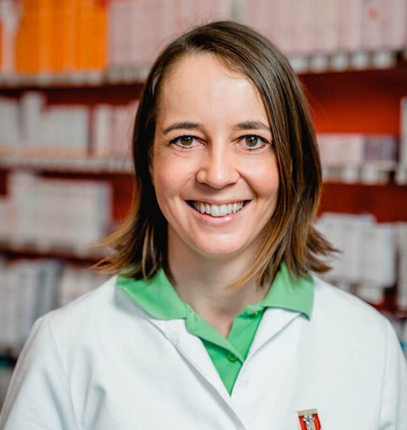 Eine Frau in einem weißen Laborkittel und einem grünen Hemd mit Kragen lächelt vor Regalen voller Schachteln in einer Apotheke oder einer medizinischen Einrichtung der Laurentius Apotheke Imst / Pitztal Apotheke.