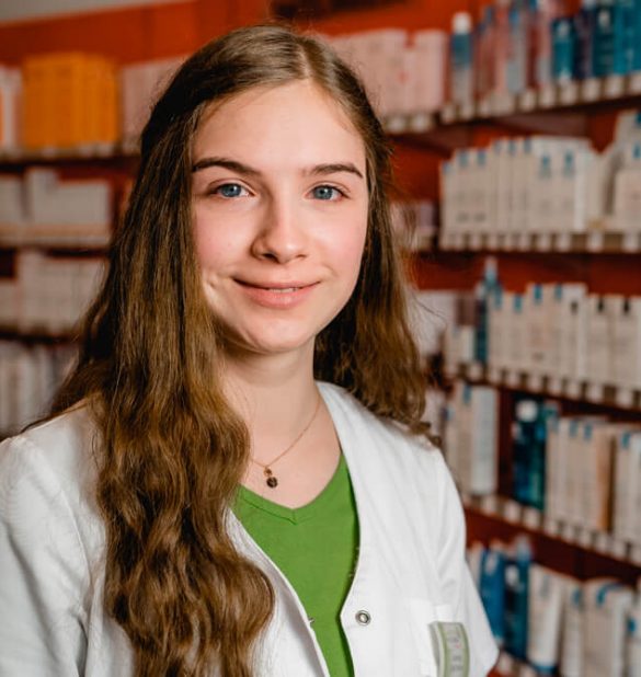 Eine junge Frau in einem weißen Kittel steht vor Regalen voller Apothekenartikel in der Laurentius Apotheke Imst / Pitztal Apotheke und lächelt in die Kamera.