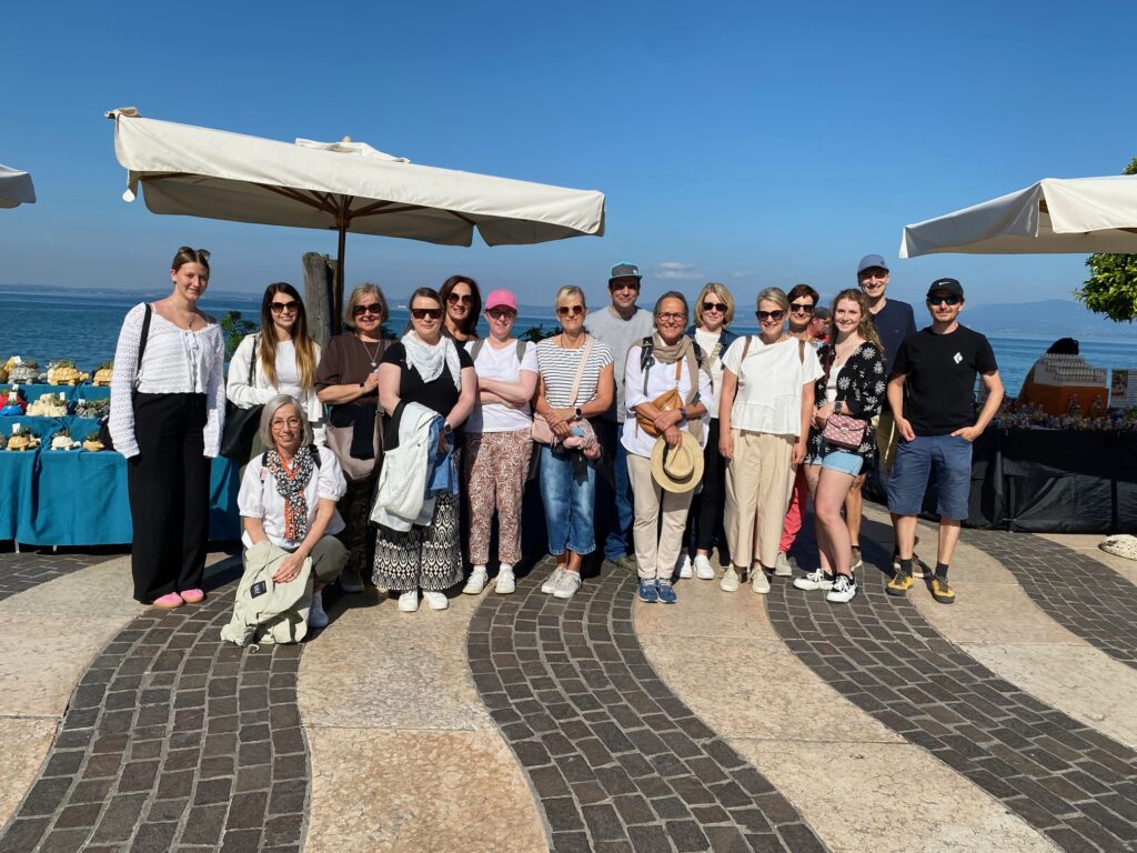 Eine Gruppe von vierzehn Erwachsenen aus der Laurentius Apotheke Imst / Pitztal Apotheke posiert für ein Foto auf einer sonnigen Promenade am Meer, vor Marktständen und großen weißen Sonnenschirmen.